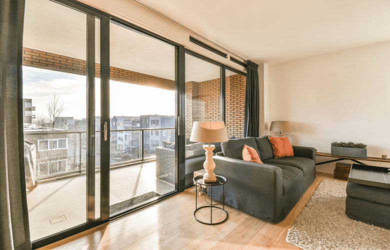 Een appartement met een gezellige living met een grijze zetel, grote zwarte schuiframen en zicht op een woonwijk.