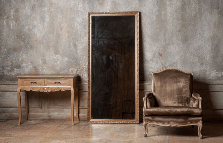 Een spiegel met donker gekleurd glas met een tafeltje en een antieke zetel voor een grijze muur.